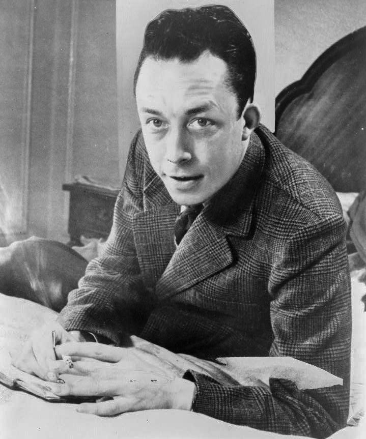 Photograph of a man with swept-back hair in a jacket and tie, leaning on a desk