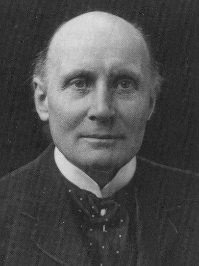 Photograph of a bald man with a wing collar and a spotted tie