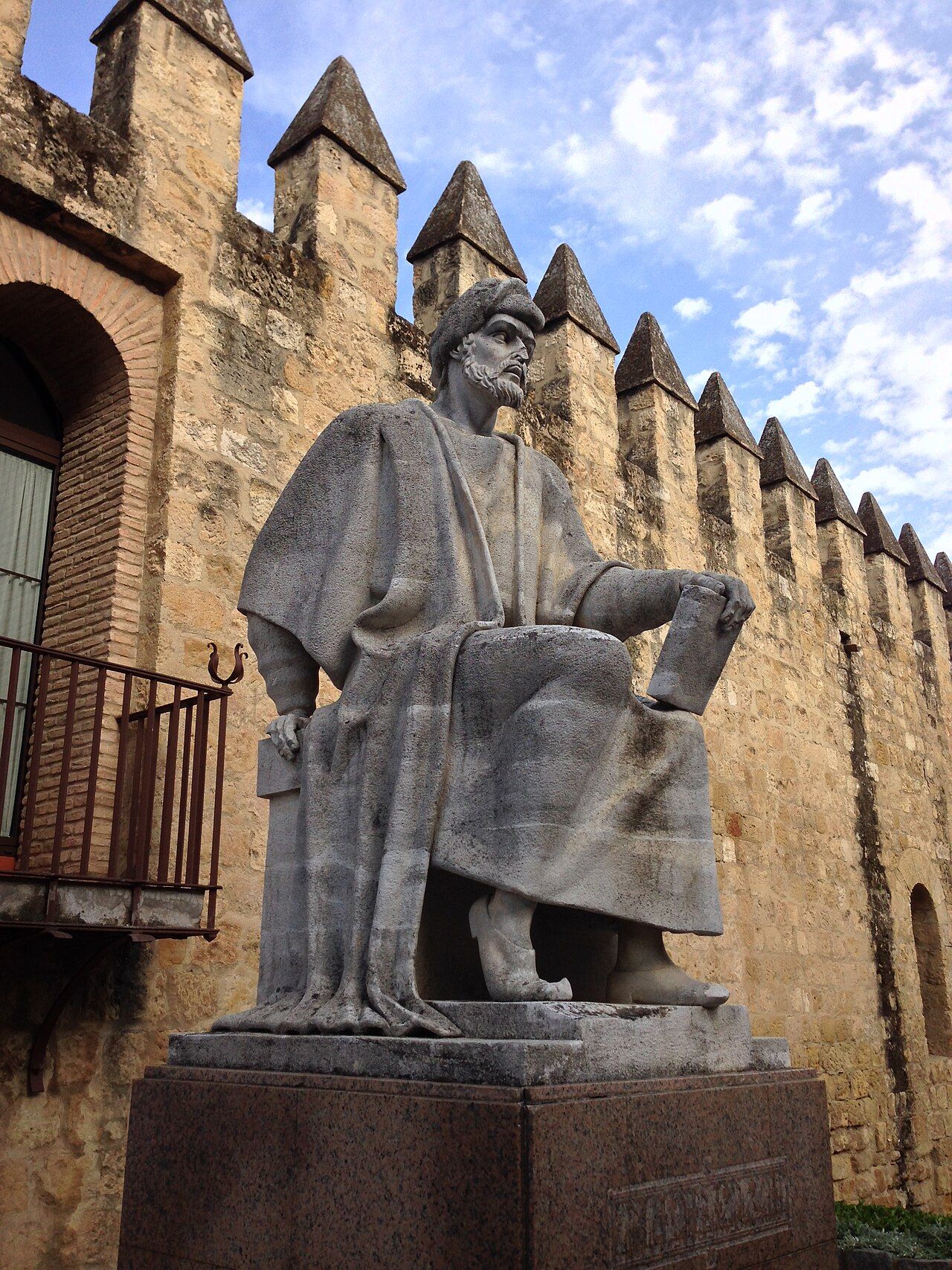 Stone statue of a seated, bearded man in a turban and robes, holding a book, beside a crenellated city wall