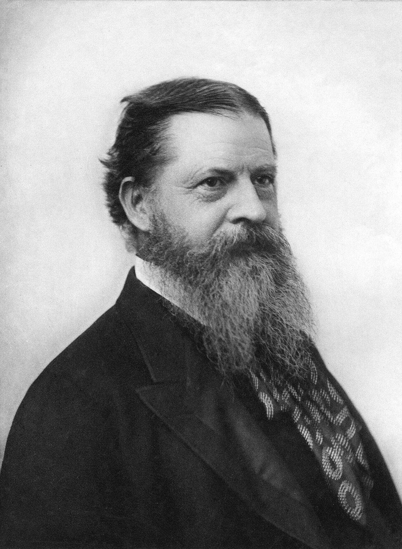Photograph of a man with a full beard and swept-back hair, in a dark coat and patterned cravat