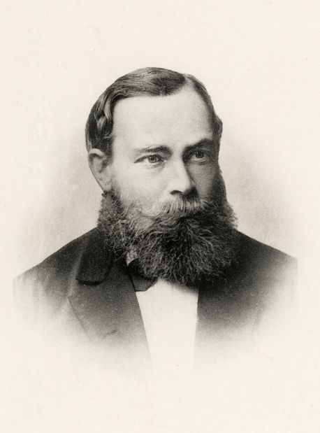 Photograph of a young man with a full beard and neatly parted hair, in a dark jacket