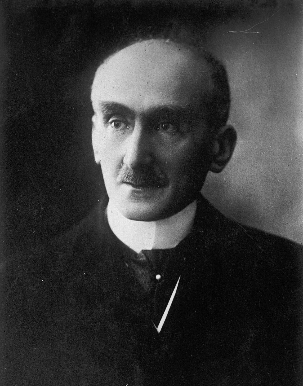 Photograph of a balding man with a moustache in a dark suit and high collar