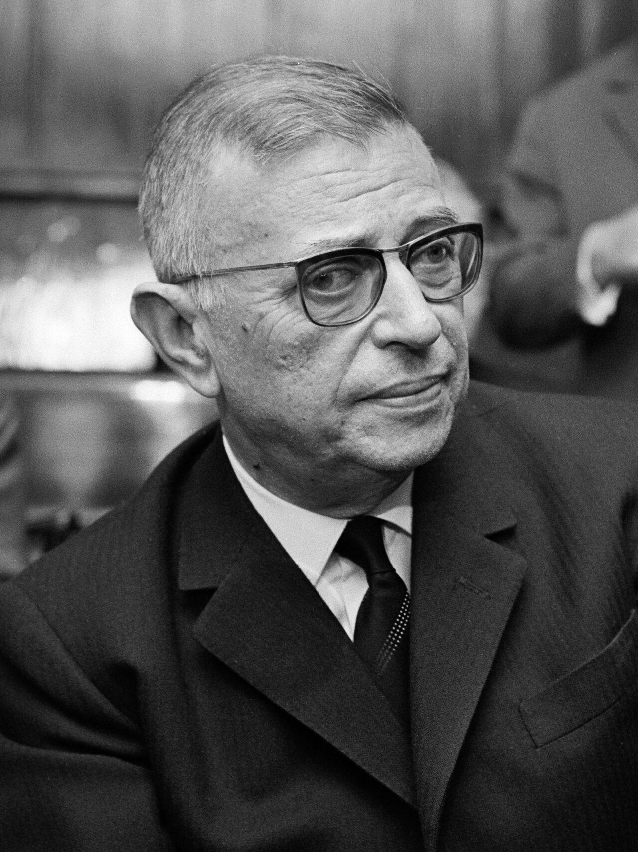 Black and white photograph of a man in thick glasses and a dark suit, looking to one side