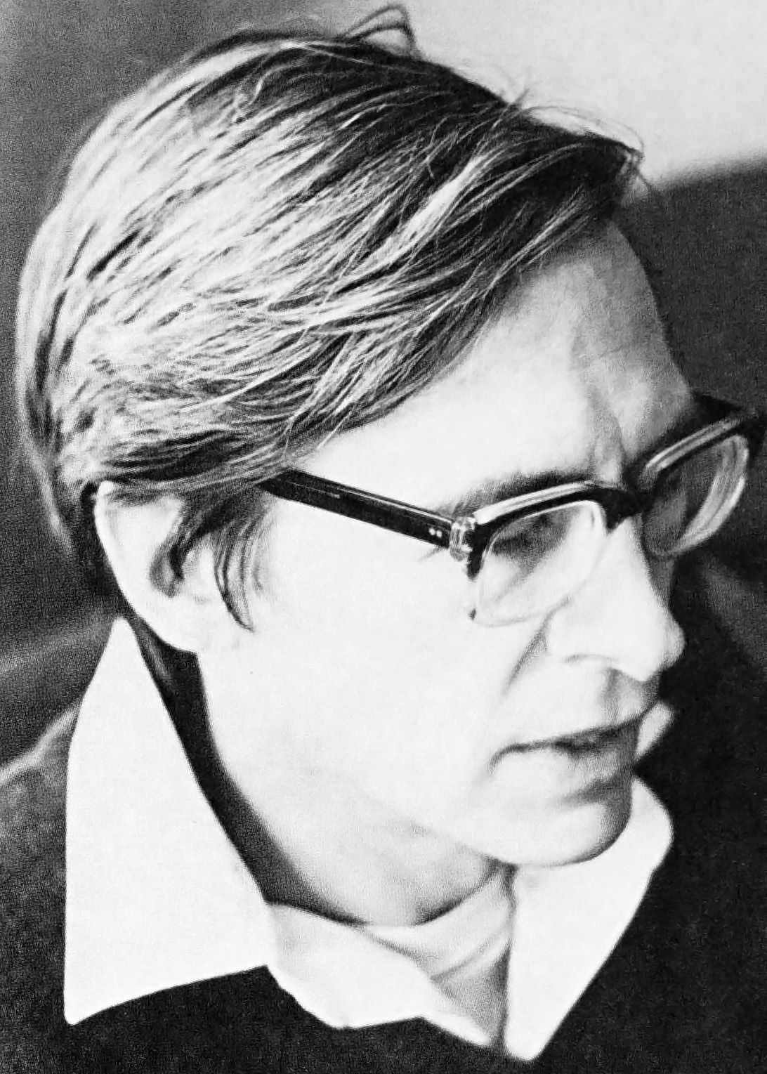 Photograph of a man with a fringe of dark hair and heavy-framed glasses, looking down