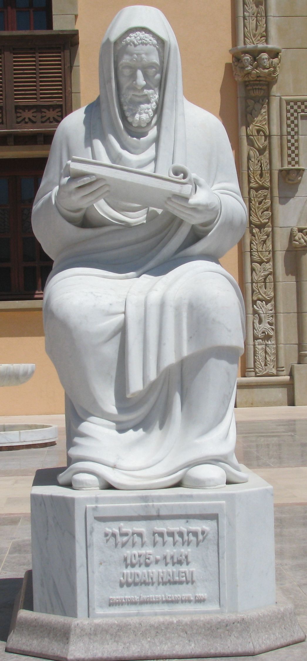 White statue of a seated, hooded man with a beard, holding a book on his lap
