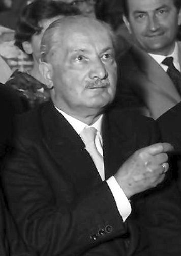 Photograph of an older man with a moustache in a dark suit at a public event