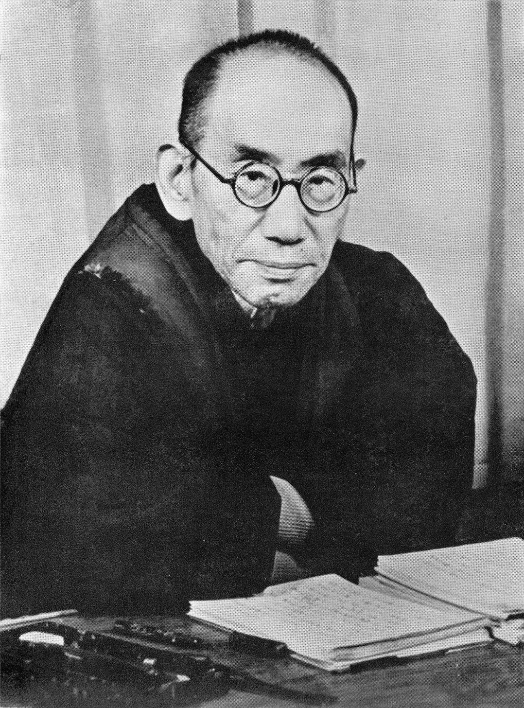 Black-and-white photograph of an elderly man in round glasses and a dark kimono, seated at a desk with open books