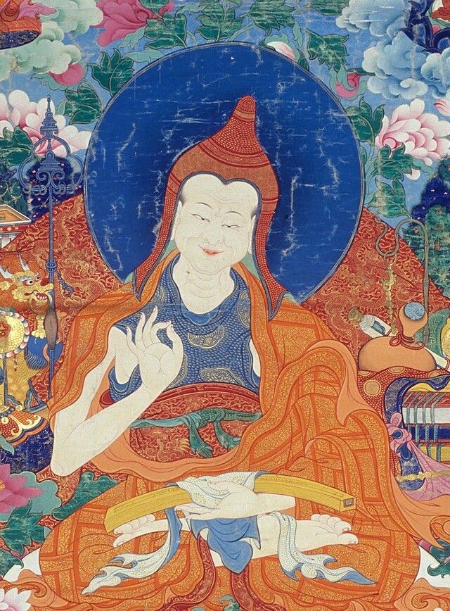 Painted figure of a smiling monk in a pointed red hat and orange robes, one hand raised, before a dark blue halo