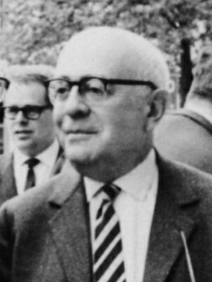 Photograph of a balding man in glasses and a striped tie, with other men behind him