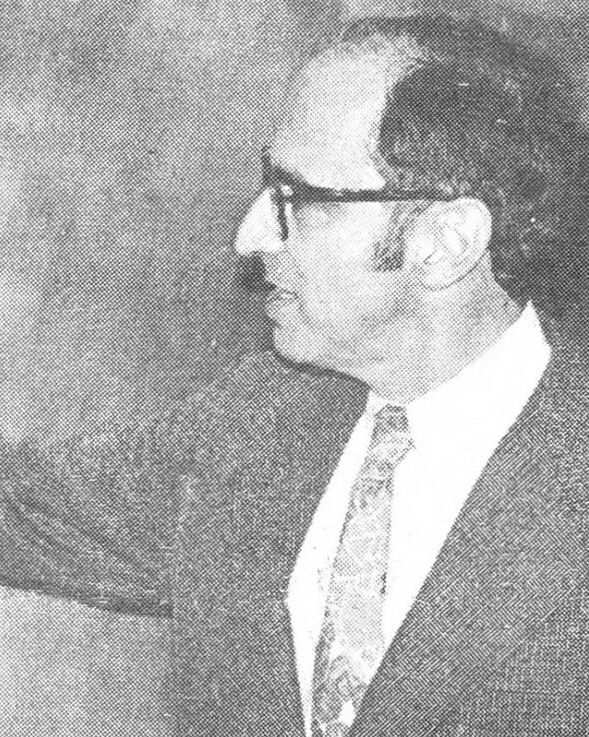 Photograph of a man in glasses and a patterned tie, in profile at a blackboard