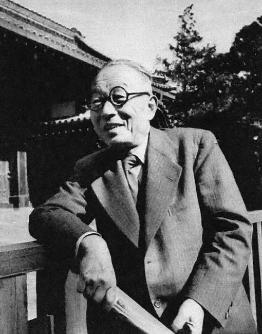 Black-and-white photograph of an elderly man in round glasses and a suit, leaning on a wooden railing